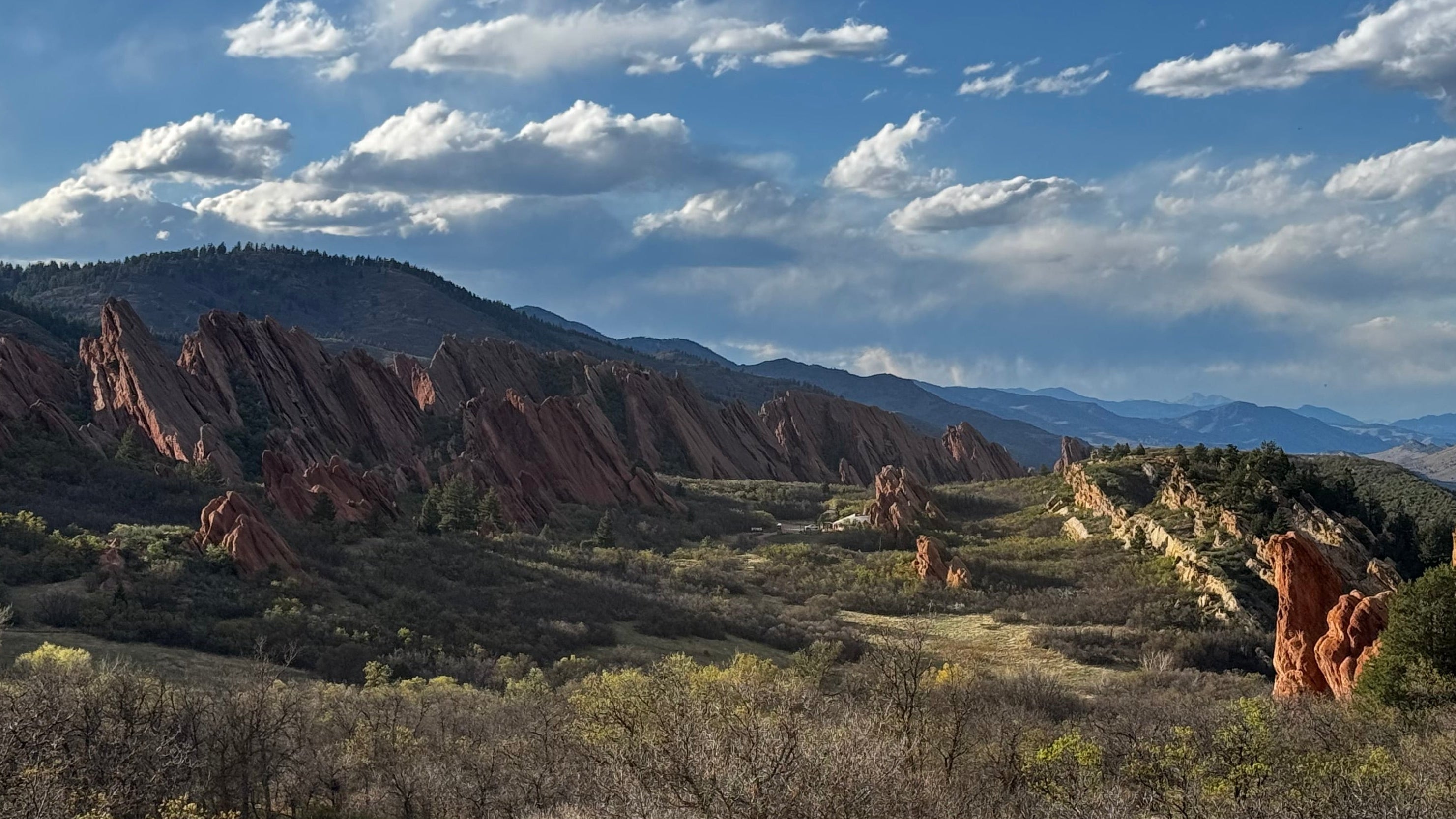 A scenic landscape photograph of red rock formations and mountains under a blue sky with white clouds, with dry grass and shrubs in the foreground.